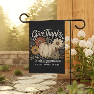 Puede incluir: Una bandera de jardín rectangular negra con una calabaza color crema en el centro rodeada de flores en tonos otoñales naranja, amarillo y verde salvia. El texto dice Give Thanks in all circumstances 1 THESSALONIANS 5:18.