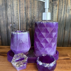 Op de afbeelding: Een paarse badkamerset met een geometrisch ontwerp. De set bevat een zeepdispenser met een zilveren pomp, twee kleine bekers, een dienblad en een zeepbakje. De randen van de bekers en het zeepbakje zijn versierd met zilveren glitter.