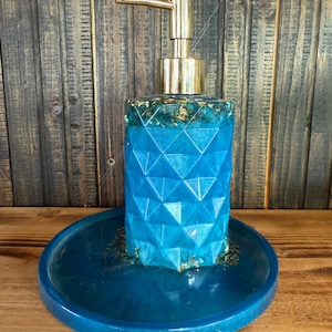 Op de afbeelding: Een turquoise blauwe zeepdispenser met een geometrisch patroon en gouden accenten. De dispenser staat op een bijpassend turquoise blauw dienblad. De pomp is goudkleurig. De achtergrond is een houten oppervlak.