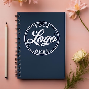 Może przedstawiać: Niebieski notes w spirali z białym kółkiem na okładce, na którym widnieje napis "YOUR LOGO HERE". Notes leży na różowym tle z kwiatami.