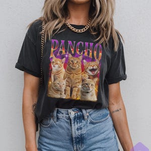 May include: Dark gray t-shirt featuring a graphic of five orange tabby cats with the word "PANCHO" in a colorful, retro font. The cats are arranged in a row, with one cat smiling.