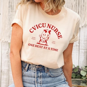 Puede incluir: Una camiseta de color crema con un gr&aacute;fico rojo que dice "CVICU NURSE" y presenta un coraz&oacute;n de dibujos animados sosteniendo un estetoscopio. El texto debajo del gr&aacute;fico dice "ONE BEAT AT A TIME".