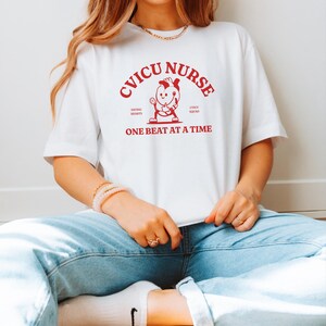 Puede incluir: Camiseta blanca con texto rojo que dice "CVICU NURSE" con un coraz&oacute;n de dibujos animados sosteniendo un estetoscopio y el texto "Saving Hearts CVICU Squad One Beat At A Time".