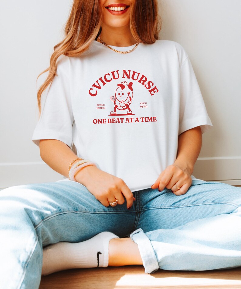 Puede incluir: Camiseta blanca con texto rojo que dice "CVICU NURSE" con un coraz&oacute;n de dibujos animados sosteniendo un estetoscopio y el texto "Saving Hearts CVICU Squad One Beat At A Time".