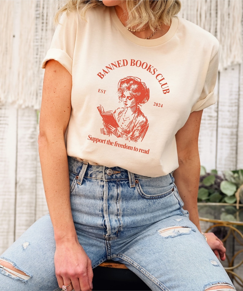 May include: A beige t-shirt with a red graphic of a woman reading a book. The graphic says "Banned Books Club Est 2024" and "Support the freedom to read".