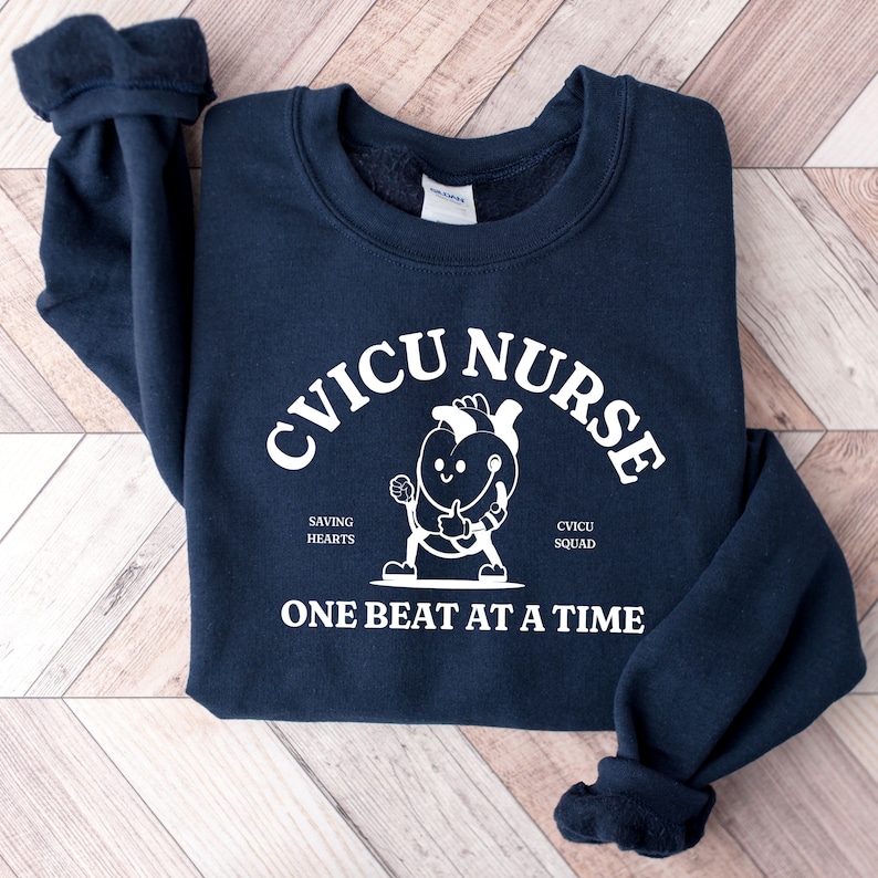 May include: Navy blue sweatshirt with a white graphic of a cartoon heart wearing a stethoscope and giving a thumbs up. The text "CVICU NURSE" is in a white arc above the heart, and the text "SAVING HEARTS" and "CVICU SQUAD" are below the heart. The text "ONE BEAT AT A TIME" is at the bottom of the graphic.