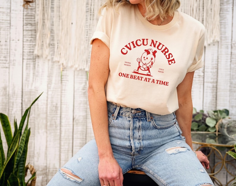 Puede incluir: Una camiseta de color crema con un gr&aacute;fico rojo que dice "CVICU NURSE" y presenta un coraz&oacute;n de dibujos animados sosteniendo un estetoscopio. El texto debajo del gr&aacute;fico dice "ONE BEAT AT A TIME".
