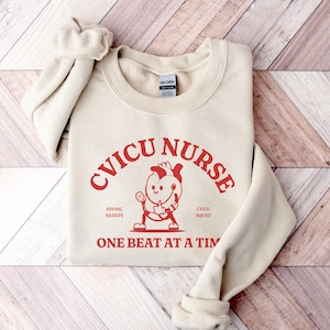 May include: A beige sweatshirt with the text "CVICU NURSE" in red. The text is in an arc shape with a cartoon heart wearing a stethoscope and holding a heart in its hand. The text "SAVING HEARTS" and "CVICU SQUAD" are below the arc. The text "ONE BEAT AT A TIME" is below the cartoon heart.