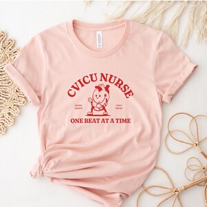 Puede incluir: Camiseta rosa con el texto "CVICU NURSE" en rojo, con una ilustraci&oacute;n de dibujos animados de una enfermera sosteniendo un estetoscopio y un coraz&oacute;n. El texto "Saving Hearts, CVICU Squad, One Beat at a Time" tambi&eacute;n est&aacute; en rojo.