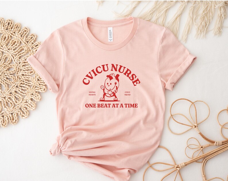 Puede incluir: Camiseta rosa con el texto "CVICU NURSE" en rojo, con una ilustraci&oacute;n de dibujos animados de una enfermera sosteniendo un estetoscopio y un coraz&oacute;n. El texto "Saving Hearts, CVICU Squad, One Beat at a Time" tambi&eacute;n est&aacute; en rojo.