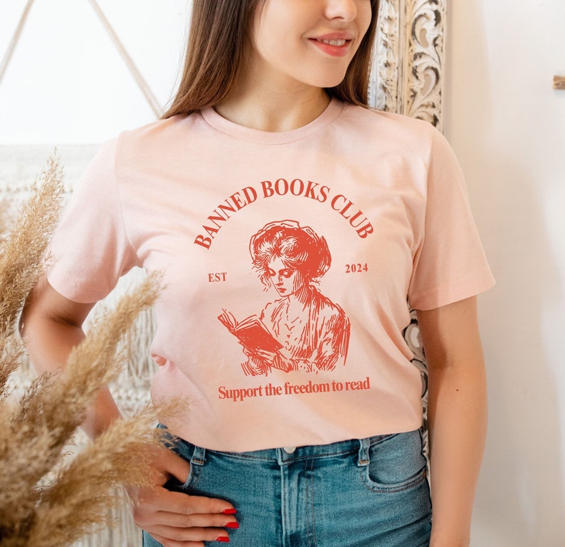 May include: A pink t-shirt with a red graphic design that says "Banned Books Club Est 2024" and features a woman reading a book. The graphic also says "Support the freedom to read".