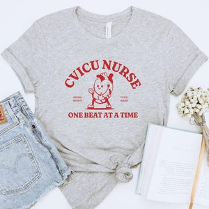 Puede incluir: Una camiseta gris con el texto "CVICU NURSE SAVING HEARTS ONE BEAT AT A TIME" en rojo. Un coraz&oacute;n de dibujos animados con cara y brazos sostiene un estetoscopio.