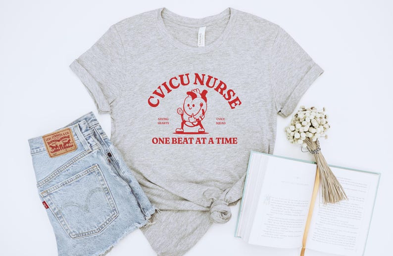 Puede incluir: Una camiseta gris con el texto "CVICU NURSE SAVING HEARTS ONE BEAT AT A TIME" en rojo. Un coraz&oacute;n de dibujos animados con cara y brazos sostiene un estetoscopio.