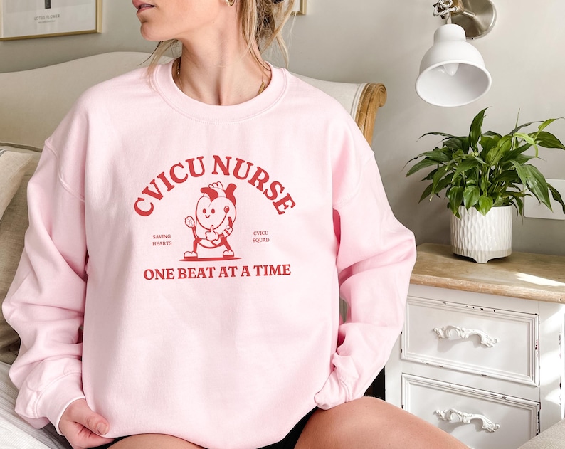 May include: Pink sweatshirt with the text "CVICU NURSE" in red, featuring a cartoon heart with a stethoscope and the text "Saving Hearts" and "CVICU Squad" on either side. The text "One Beat At A Time" is below the cartoon heart.