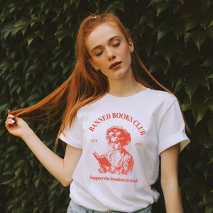 May include: White t-shirt with a red graphic design that says "Banned Books Club" with an illustration of a woman reading a book. The text also says "Est 2024" and "Support the freedom to read".