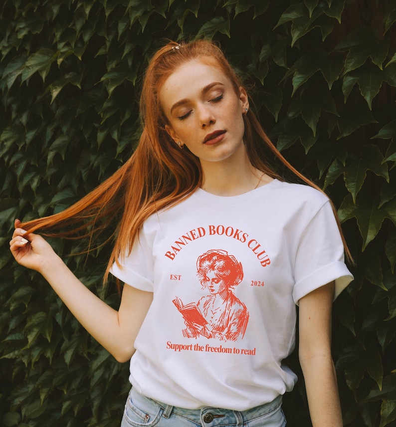 May include: White t-shirt with a red graphic design that says "Banned Books Club" with an illustration of a woman reading a book. The text also says "Est 2024" and "Support the freedom to read".