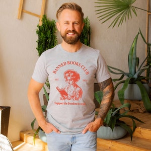 May include: A grey t-shirt with a red graphic of a woman reading a book. The graphic says "Banned Books Club Est 2024" and "Support the freedom to read".