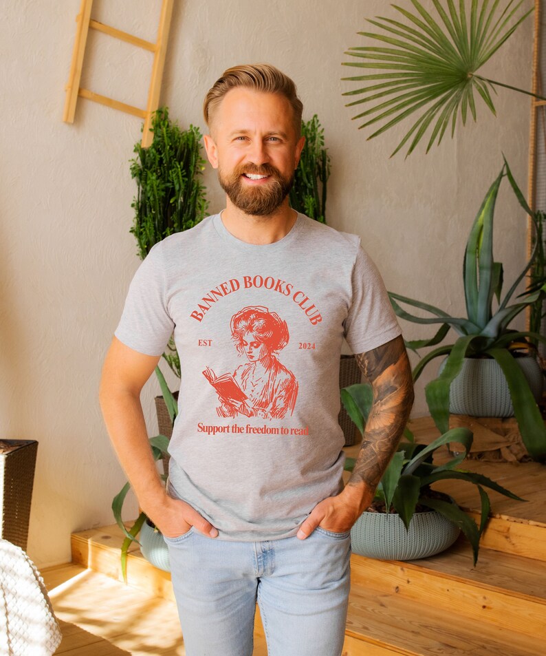 May include: A grey t-shirt with a red graphic of a woman reading a book. The graphic says "Banned Books Club Est 2024" and "Support the freedom to read".