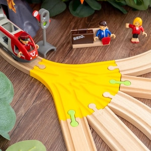 Puede incluir: Un juego de tren de juguete de madera con un tren rojo y blanco, vías y figuras en miniatura. El juego incluye un conector de vía amarillo, una señal y una estación con la palabra "BRIO". El tren entra en un túnel gris.