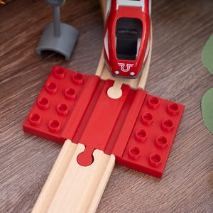 Könnte beinhalten: Ein roter Spielzeugzug mit weißem Streifen und ein rotes Brückenteil mit puzzleartiger Verbindung. Der Zug befindet sich auf einer Holzstrecke. Ein graues Spielzeugsignal befindet sich im Hintergrund. Die Szene spielt auf einer Holzoberfläche.