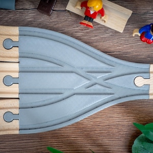 May include: Wooden toy train tracks with a gray switch track, a red toy car, and two toy figures. The tracks are on a brown wooden surface, with green leaves and small toy accessories scattered around.