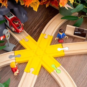 Può includere: Set di trenini giocattolo in legno con un treno rosso e grigio, un'intersezione di binari gialla e figure in miniatura. Include un segnale, una scatola marrone con la scritta "BRIO" e fogliame decorativo.