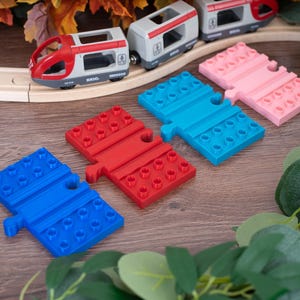 Op de afbeelding: Een Brio speelgoedtreinset met een grijze en rode locomotief en passagierswagon op een houten rails, vergezeld van drie rechthoekige plastic railadapters in blauw, rood en lichtblauw, en een roze adapter, elk voorzien van bouwsteennopjes.