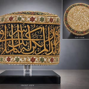 Op de afbeelding: Een cilindervormige muts van zwarte stof met goudkleurig geborduurde Arabische kalligrafie. De boven- en onderrand zijn versierd met een crèmekleurige band met rode en groene bloemmotieven. Het bovenaanzicht toont een cirkelvormig crèmekleurig oppervlak met gedetailleerd goudborduursel en een bloemenrand.