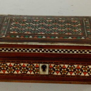 May include: A rectangular wooden box with intricate inlaid designs. The lid and front feature geometric patterns in red, green, and white against a dark brown background. The box has a keyhole and is trimmed with a checkered pattern.