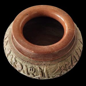 Puede incluir: Una maceta de terracota con una abertura ancha y redondeada y tallas intrincadas alrededor del borde. La maceta está decorada con un patrón de figuras estilizadas y diseños geométricos.