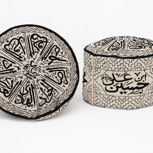 Könnte beinhalten: Ein Paar runde, cremeweiße Kufi-Kappen mit aufwendiger schwarzer arabischer Kalligrafie-Stickerei und geometrischen Mustern. Die Oberseite einer Kappe zeigt ein radiales Design, während die Seite der anderen kräftige Schriftzeichen und einen Zickzackrand aufweist.