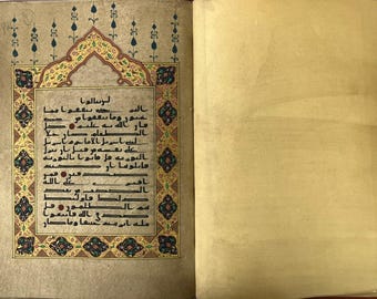 May include: An open book with a beige page on the right and a decorated page on the left. The left page features black Arabic calligraphy centred within a rectangular frame, surrounded by a gold, blue, and red floral border with pointed arches.