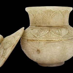 Puede incluir: Jarra tallada de color marfil con tapa. La jarra tiene un cuerpo redondeado y un borde acampanado, con intrincados diseños florales y de hojas. La tapa tiene forma de hoja estilizada con un pequeño asa redondeada. Fondo negro.