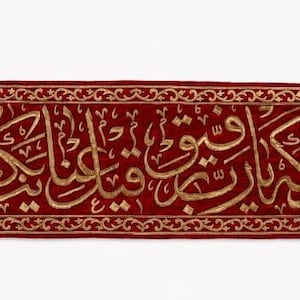 May include: A long, rectangular, deep red velvet banner featuring intricate gold-coloured embroidery of Arabic calligraphy. The banner has pointed ends, each finished with a small, light-coloured decorative tassel, and a matching gold border.