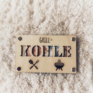 Könnte beinhalten: Ein Holzschild mit dem Text "Grill - Kohlie" in einer dekorativen Schriftart. Das Schild zeigt auch ein Grill- und Spatel-Symbol.