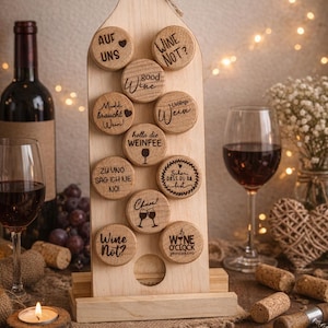 Könnte beinhalten: Ein hölzerner Wein-Flaschenförmiger Display-Ständer mit Korken mit verschiedenen Phrasen wie "Wine Not?" und "Good Wine". Der Ständer hält mehrere Korken mit graviertem Text und ist mit einer Schnur-Schleife verziert. Weingläser und eine Weinflasche sind im Hintergrund.