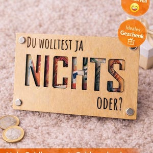 May include: A wooden gift card with the word "NICHTS" cut out, revealing money. The card reads "DU WOLLTEST JA NICHTS ODER?" and includes the Etsy logo. Additional text includes "Holz Geldkarte mit Geldgeschenk" and "SVG-Datei".