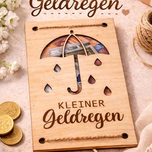 Puede incluir: Tarjeta de regalo de madera con diseño de paraguas, llena de dinero. El texto "Kleiner Geldregen" está grabado en la madera. Incluye hilo decorativo y detalles de monedas.