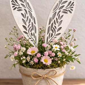 Könnte beinhalten: Osterarrangement in einem Keramiktopf mit rosa und weißen Blumen, grünem Moos und hölzernen Hasenohren mit filigranen Ausschnitten. Der Topf ist mit einer Schnur-Schleife gebunden.