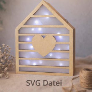 May include: A wooden house-shaped decorative light with a heart cutout. The light has horizontal slats and is illuminated by small white lights. The text "SVG Datei" is visible at the bottom.