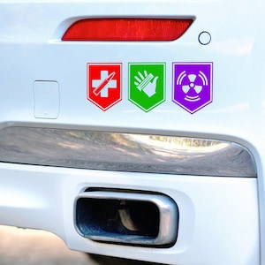 May include: Three colourful vinyl decals on a white car bumper. The decals are shield-shaped with white graphics: a red cross and bullet, a green hand with a glowing effect, and a purple radiation symbol.