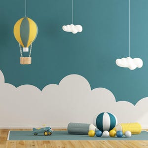Puede incluir: Una escena de guardería caprichosa con un globo aerostático amarillo y gris, decoraciones en forma de nube y una pelota a rayas azul y blanca. La habitación tiene una pared verde azulado con recortes de nubes blancas y una alfombra azul.