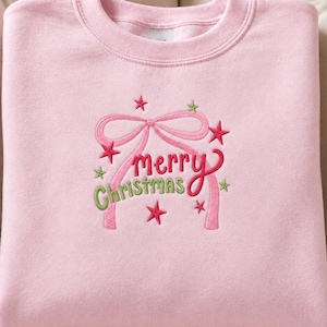 May include: A light pink crewneck sweatshirt featuring an embroidered design of a pink ribbon bow, small red and green stars, and the text "merry Christmas" in red and green script lettering centered on the front of the garment.