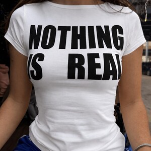 May include: A white short-sleeved t-shirt featuring the bold black text "NOTHING IS REAL" printed across the chest. The garment has a crew neckline and a fitted silhouette, constructed from a plain white fabric with high-contrast lettering.