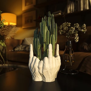 Könnte beinhalten: Ein weißer Keramik-Pflanztopf in Form von zwei Händen, die den Mittelfinger zeigen, mit einem grünen Kaktus. Der Pflanztopf steht auf einem schwarzen Tisch, mit einer Lampe und einer Vase mit Blumen im Hintergrund.