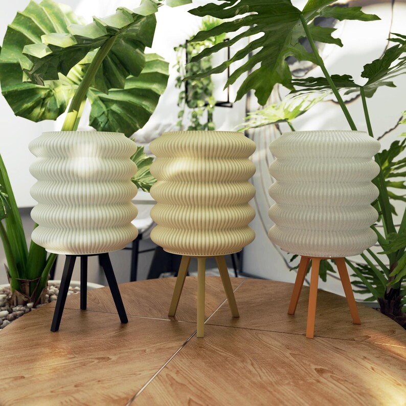 K&ouml;nnte beinhalten: Drei dekorative Pflanzgef&auml;&szlig;e mit gerippten, zylindrischen K&ouml;rpern und Dreibein-Beinen. Die Pflanzgef&auml;&szlig;e sind cremefarben, beige und wei&szlig;. Die Beine sind schwarz, beige und orange. Die Pflanzgef&auml;&szlig;e werden auf einer Holzoberfl&auml;che pr&auml;sentiert, umgeben von gr&uuml;nen Pflanzen.