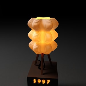 Puede incluir: Una lámpara iluminada de color crema con un diseño único y en capas. La lámpara se asienta sobre una base de madera con tres patas y un soporte rectangular. El año "1997" se muestra en naranja en la base. La lámpara emite un brillo cálido.