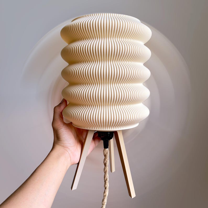 K&ouml;nnte beinhalten: Cremefarbene, gerippte Lampe mit einem einzigartigen, geschichteten Design. Die Lampe steht auf einem hellen Holzstativ und hat ein geflochtenes Kabel. Der Lampenschirm hat ein strukturiertes, wellenf&ouml;rmiges Aussehen. Das Gesamtdesign ist modern und minimalistisch.