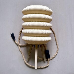 K&ouml;nnte beinhalten: Cremefarbene Tischlampe mit geripptem, zylindrischem Schirm. Die Lampe steht auf einem Dreibein und hat ein geflochtenes Seil. Ein schwarzer Stecker und ein Ein-/Ausschalter sind sichtbar. Dekorative Leuchte.