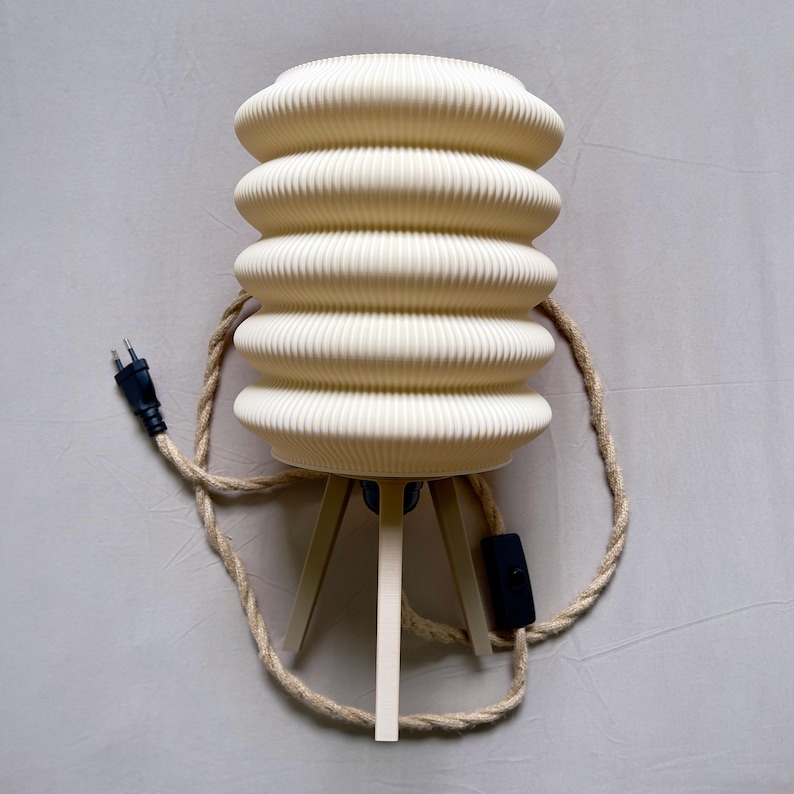 K&ouml;nnte beinhalten: Cremefarbene Tischlampe mit geripptem, zylindrischem Schirm. Die Lampe steht auf einem Dreibein und hat ein geflochtenes Seil. Ein schwarzer Stecker und ein Ein-/Ausschalter sind sichtbar. Dekorative Leuchte.